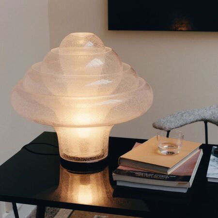 Glas als Kunst. Licht als Stimmung.  Die Lotus Tischleuchte von Carlo Nason – 1969 entworfen und nach mehr als 50 Jahren von GUBI neu aufgelegt – ist ein Meisterwerk der Murano-Glaskunst. Vier ineinander verschachtelte, mundgeblasene Glasschirme bilden eine konzentrische, blütenartige Struktur, die schon ohne Licht endlos fasziniert. Hergestellt mit der historischen «Pulegoso»-Technik aus den 1920er-Jahren, bei der winzige Luftblasen ins Glas geblasen werden, erhält jedes Stück eine halbtransparente Textur, die Tiefe und Lichtstreuung auf einzigartige Weise verstärkt – und jede Leuchte zum Unikat macht.  Eingeschaltet erzeugt die Lotus ein sanftes, diffuses Leuchten ganz ohne Blendung oder sichtbare Lichtquelle – pure Atmosphäre, die jedem Raum Ruhe und Wärme verleiht. Nur die erfahrensten Murano-Glasbläser sind in der Lage, Stücke dieser Grösse und Komplexität zu schaffen. Das Ergebnis ist eine Leuchte, die weit mehr ist als Beleuchtung – sie ist ein Kunstwerk.
